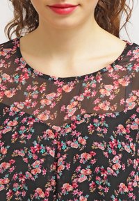 Blouse noire transparente avec un motif floral en rose, orange et vert. Présente un col rond et une coupe légèrement ample.