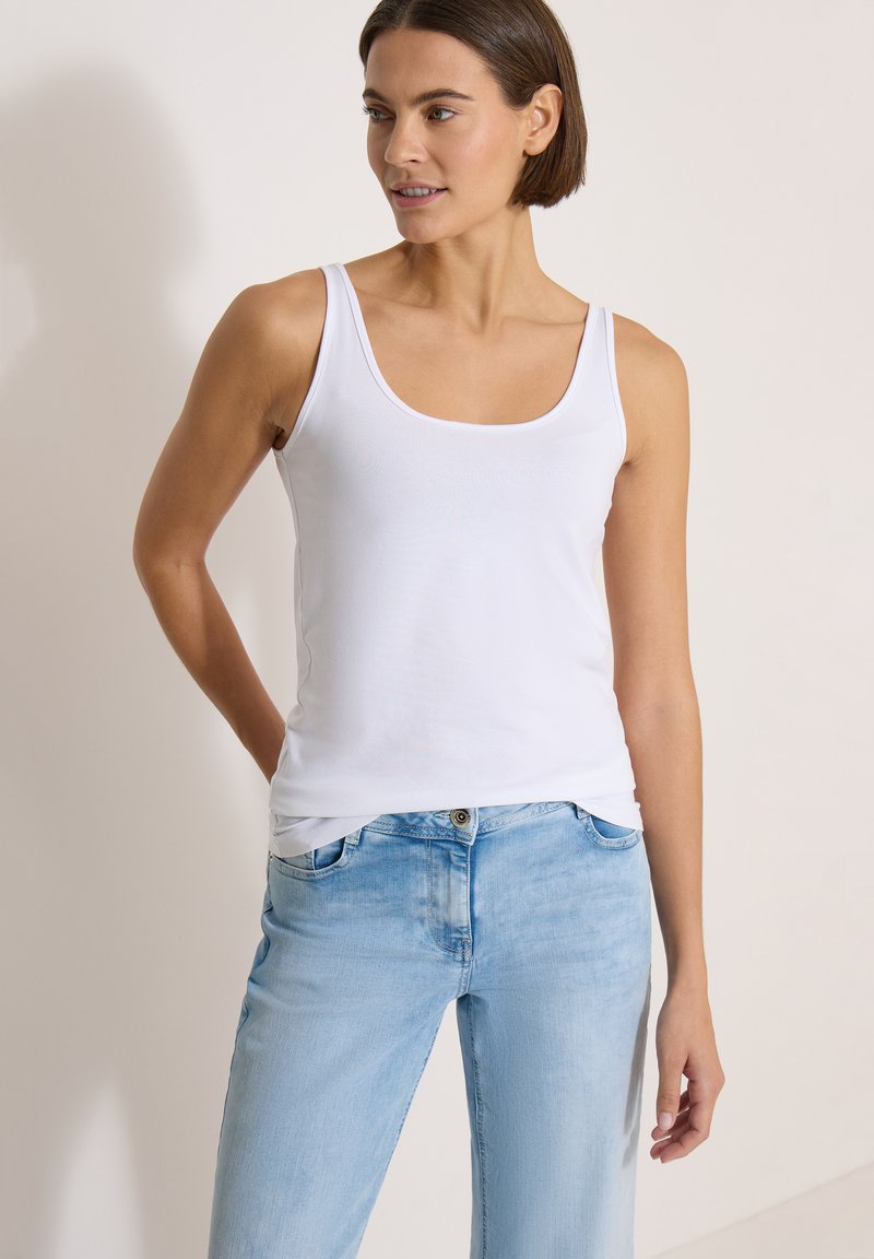Femme portant un débardeur blanc sans manches et un jean bleu clair, debout avec une main dans le dos, regardant sur le côté.