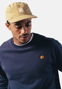 Sudadera azul marino con un logo de elefante naranja bordado en el pecho; gorra beis con un parche que presenta el mismo logo.