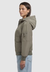 Veste légère de couleur vert olive avec capuche, devant zippé, poignets côtelés et poche poitrine. Tissu lisse et légèrement brillant.