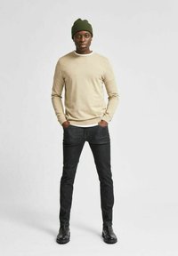 Camicia beige a maniche lunghe con colletto bianco, jeans scuri e stivali neri. Il modello indossa un berretto a coste verde. In piedi all'interno, sfondo neutro.