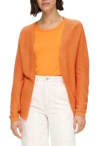 Orange gestrickte Strickjacke mit offenem Front, strukturiertem Stoff und gerippten Bündchen, getragen über einem passenden orangefarbenen Oberteil, kombiniert mit hellen, hoch taillierten Hosen.