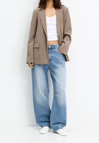 Blazer beige oversized par-dessus un débardeur blanc ajusté, associé à un jean large bleu clair avec des détails de couture visibles et des baskets blanches.
