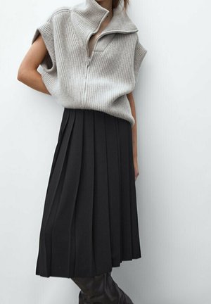Femme portant un pull beige côtelé sans manches avec fermeture éclair, associée à une jupe midi noire plissée et des bottes noires sur un fond blanc uni.
