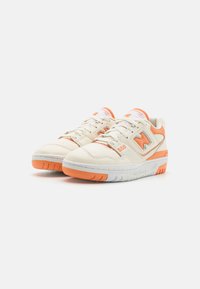 O pereche de adidași New Balance 550, albi și portocalii, cu șireturi, branding și tălpi din cauciuc portocalii, pe un fundal alb.
