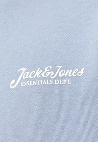 Jasnoniebieska tkanina o gładkiej fakturze, z białym haftowanym tekstem "Jack & Jones Essentials Dept." wyraźnie wyeksponowanym.