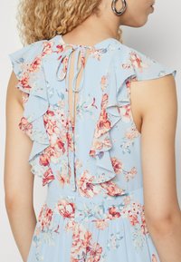 Ανθοστόλιστο φόρεμα από ανοιχτό μπλε ύφασμα με επιπέδια ruffles κατά μήκος της λαιμόκοψης και της πλάτης, διαθέτοντας κλείσιμο με κορδόνι και παστέλ λουλουδάτα σχέδια.