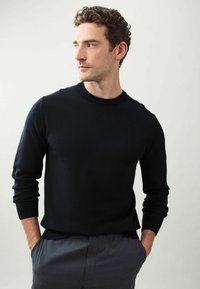 Pulover negru din tricot cu decolteu rotund, mâneci și tiv cu nervuri. Asortat cu pantaloni gri, modelul stă pe un fundal neutru.