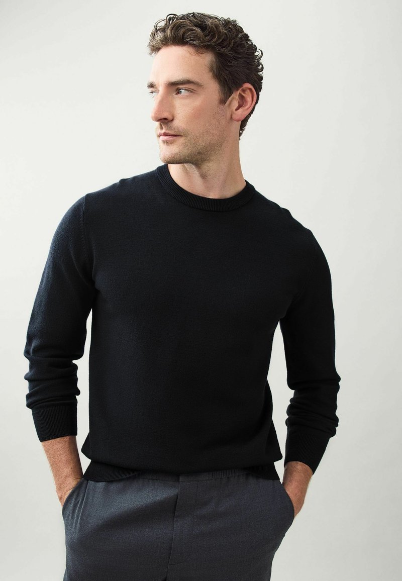 Pulover negru din tricot cu decolteu rotund, mâneci și tiv cu nervuri. Asortat cu pantaloni gri, modelul stă pe un fundal neutru.