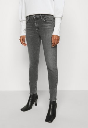 Personne portant un jean gris ajusté, des bottines noires à talons hauts et un haut blanc à manches longues, debout devant un fond clair uni.