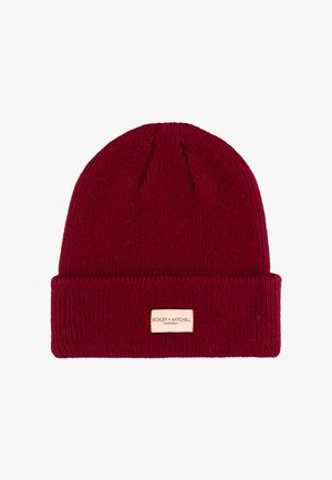 Deep red knit beanie with a folded cuff, featuring a small rectangular brand label in beige stitched on the front.