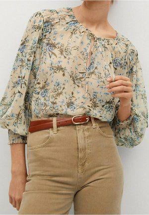 Blouse florale crème avec des fleurs bleues, manches bouffantes, et un col en forme de clé. Associée à un pantalon beige taille haute et une ceinture marron.