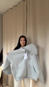 Felpa grigia con zip realizzata in tessuto morbido, dotata di un grande cappuccio, tasche a marsupio e etichetta attaccata. Texture liscia con maniche ampie.