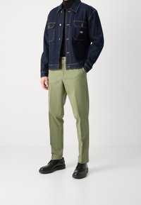 Giubbotto in denim blu scuro con cuciture gialle, abbinato a pantaloni chinos verde chiaro e scarpe in pelle nere, creando un look casual a strati.