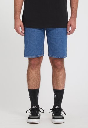 Hombre de pie que lleva puestos pantalones cortos de mezclilla azules, calcetines negros a media pantorrilla con logotipos blancos, zapatillas negras y grises, y una camiseta negra.