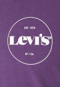 Logo Levi's en blanc avec le texte "EST. 1873" et "SF / CA." à l'intérieur d'un cercle sur fond en tissu violet.