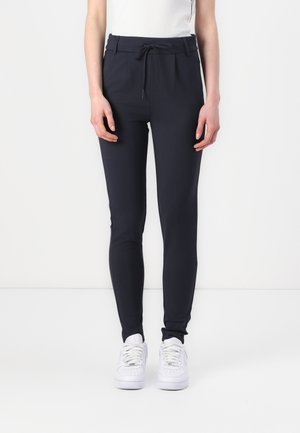 Persoon die zwarte slim-fit broek met trekkoord in de taille en witte sneakers draagt, staand tegen een effen witte achtergrond.