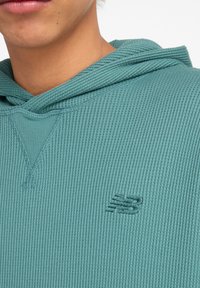 Sweat à capuche côtelé bleu sarcelle avec col en V et logo brodé sur la poitrine. Présente un tissu texturé et une capuche avec cordon de serrage.