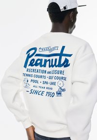 Witte sweatshirt met blauwe bedrukte tekst en illustraties. Tekst bevat "Peanuts" en activiteiten zoals tennis, golf en biljart sinds 1950.