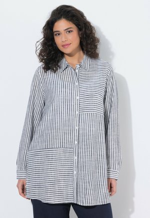 Kvinde med krøllet hår iført en lang, sort-hvid stribet skjorte med knapper og varierende striberetninger, stående mod en hvid baggrund.