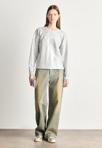 Mulher vestindo um sweatshirt de lantejoulas prateadas, jeans largos desbotados, tênis prateados e segurando uma pequena clutch prateada contra um fundo simples.