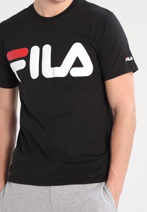 Uomo che indossa una t-shirt FILA nera con grande logo bianco e rosso sul petto e logo più piccolo sulla manica sinistra, mani nelle tasche.