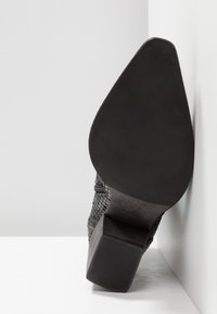 Chaussure à talon noir avec une surface texturée, dotée d'un bout pointu et d'un talon bloc. Le dessous a une finition lisse avec une légère texture mate.