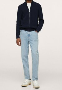 Erwachsener trägt einen navyblauen Reißverschlusspullover, helle Jeans und weiße Sneakers mit beigen Sohlen vor einem einfarbigen Hintergrund.