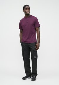 T-shirt castanho com mangas curtas, decote redondo e pequeno detalhe de logótipo. Calças cargo pretas com bolsos, usadas com ténis em preto e branco.