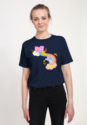 Donna che indossa una maglietta blu navy con orsi rosa e viola che giocano tra le nuvole e un arcobaleno, circondata da stelle gialle.
