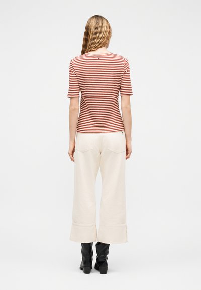 Γυναίκα με κυματιστά μαλλιά, φορώντας μπλουζάκι με κόκκινες και λευκές ρίγες και κοντά μανίκια, κρεμ παντελόνι με φαρδιές φούστες και μαύρα μποτάκια αστραγάλου, που στέκεται στραμμένη προς τα πίσω.