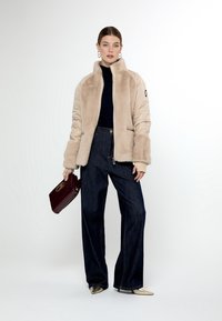 Giacca in pelliccia sintetica beige e gonfia con colletto alto, abbinata a jeans a gamba larga scuri e tacchi a punta oro. Porta una pochette bordeaux.