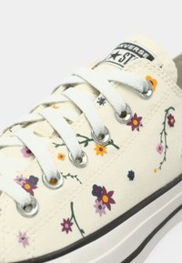 Cream canvas sneaker with white laces, silver eyelets, multicolored floral print, and a visible Converse logo on the tongue.