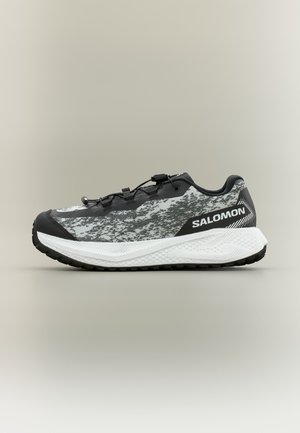 Zapatilla de running Salomon en blanco y negro con parte superior con patrón, suela blanca texturizada y sistema de cordones rápidos sobre un fondo neutro.