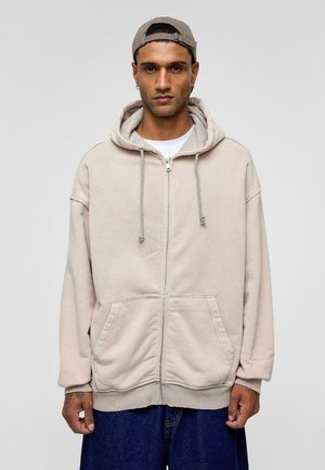 Lichtbeige zip-up hoodie met een capuchon met trekkoord, voorzakken en geribde boorden. Draag het over een wit T-shirt, gecombineerd met blauwe jeans.