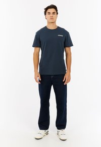 Marineblauw katoenen t-shirt met korte mouwen, ronde hals en logo aan de voorkant; gecombineerd met donkerblauwe jeans en witte sneakers.