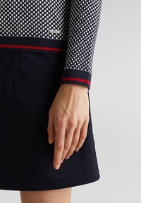 Niebieski sweter w kratę z prążkowanymi mankietami, ozdobiony czerwonymi paskami. Noszony z jednolitą granatową spódnicą, na której widać dłoń, spoczywającą na materiale.