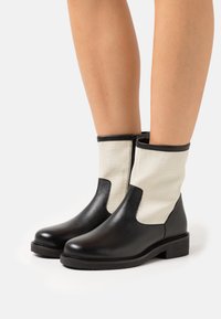 Botas de tornozelo de couro preto com laterais de lona bege, com um acabamento liso, ponta arredondada e um salto bloco baixo. Fecho com zipper de um lado.