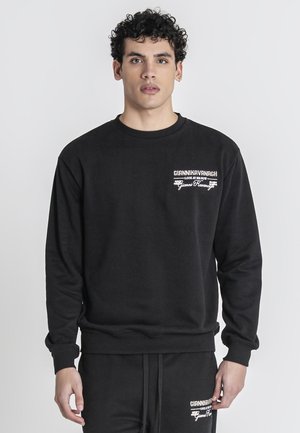 Joven con cabello rizado que lleva sudadera negra y pantalones a juego con logotipos blancos de "Gianni Kavanagh" en el pecho y el muslo.