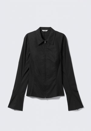 Melna pieguļoša krekla ar garām piedurknēm, smailu apkakli, priekšā rāvējslēdzēju un izvērstām aprocēm, noklāta uz gaiša fona.