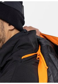 Giacca nera con accenti arancioni e dettagli con zip, caratterizzata da un alto colletto e un cappuccio regolabile. Il materiale sembra robusto e resistente alle intemperie.