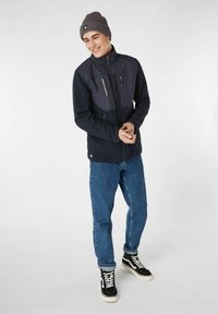 Giacca in pile navy con tasche con zip, texture a coste sulle maniche, abbinata a jeans di denim blu e scarpe alte nere. Berretto lavorato a maglia grigio.