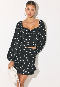 Femme portant un crop top noir à manches longues à pois et une mini-jupe à volants assortie, tenant une pochette blanche.