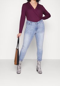 Top burdeos de manga larga cruzado, jeans ajustados de color azul claro y botas de tacón con estampado de cebra en blanco y negro. Sosteniendo un bolso de mano marrón y negro.