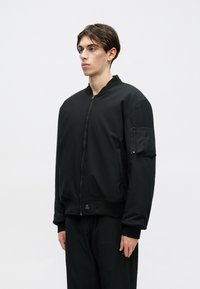 Melna bumbieru jakas mīksta, gluda materiāla ar rāvējslēdzēju priekšpusē, ribota apakšdaļa un sānu kabata kreisajā piedurknē.