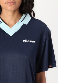 Tumši zils polo krekls ar gaiši zilu apkakli, kas veidota V veida kakla izgriezumā, zīmola "ellesse" baltā krāsā un svītrām apkakles malās.