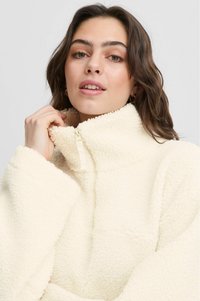 Krämfarvet fleece-pullover med høj krave, halv lynlås og blød tekstur, med brede ærmer og en boxy silhuet.