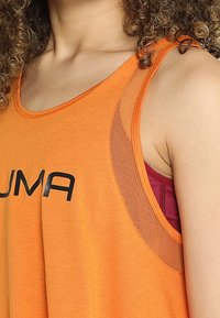 Canotta atletica arancione senza maniche con logo nero. Presenta un pannello in rete sulla spalla, sovrapposto a un reggiseno sportivo bordeaux. Tessuto morbido.