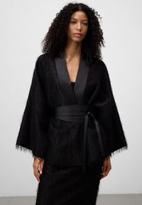 Czarny płaszcz o teksturze futra z szerokimi rękawami i satynowym paskiem. Posiada głęboki dekolt w serek oraz miękki, lekki materiał.