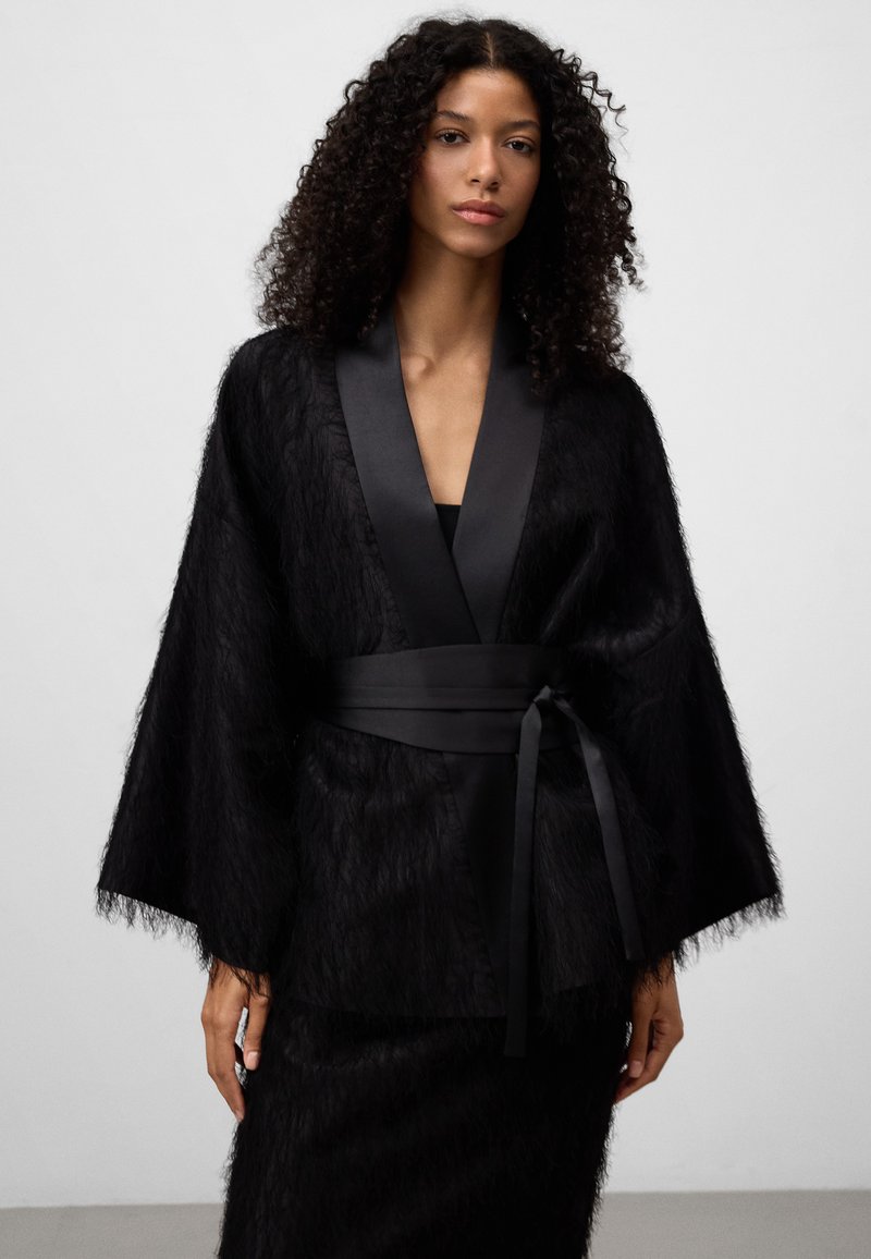 Czarny płaszcz o teksturze futra z szerokimi rękawami i satynowym paskiem. Posiada głęboki dekolt w serek oraz miękki, lekki materiał.